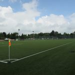 Campo de futebol, focado na linha de fundo e no escanteio, com bandeira amarela e laranja em destaque